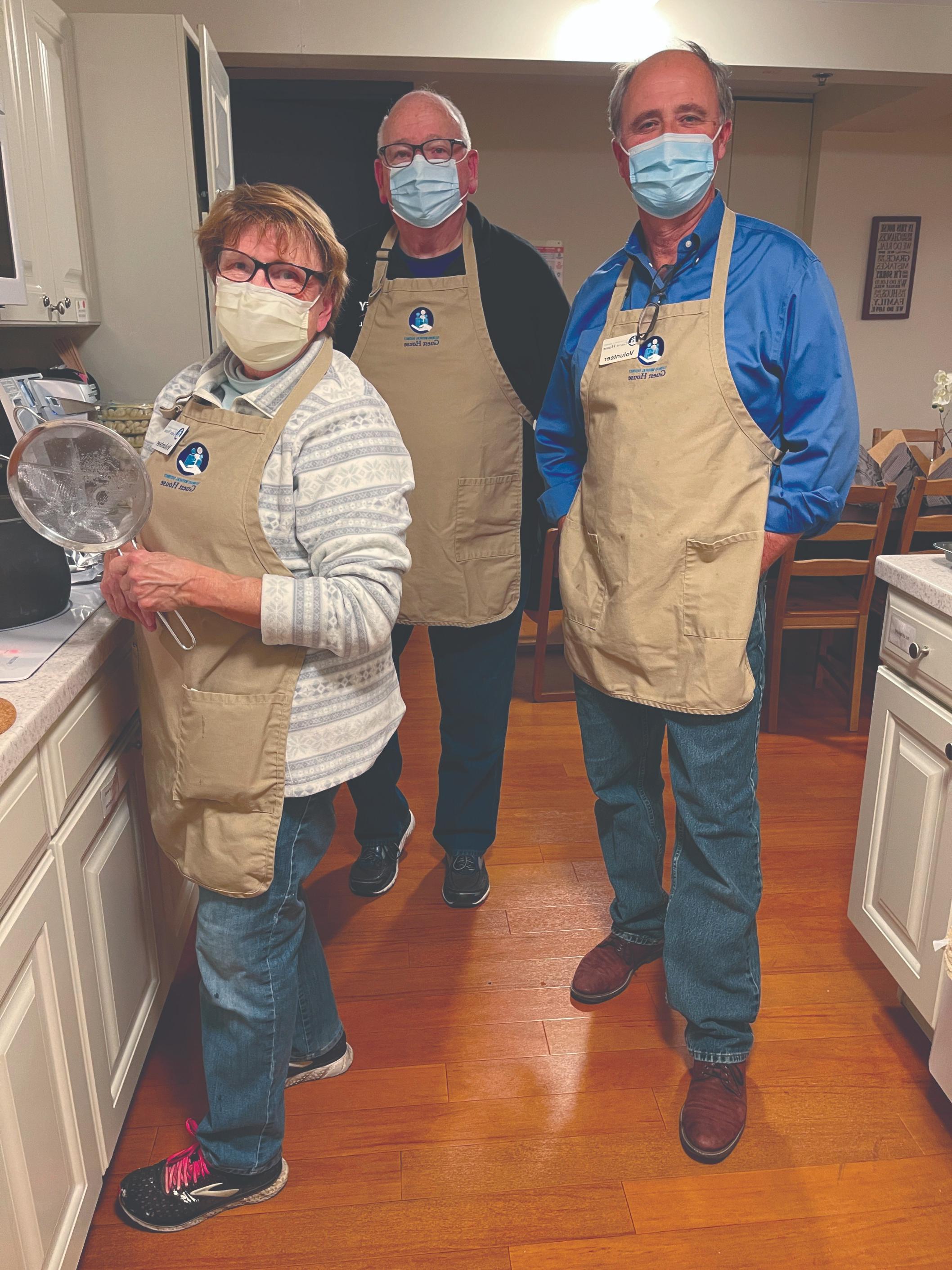 三名志愿者站在厨房里，戴着口罩，围着绣有宾馆标志的米色围裙.